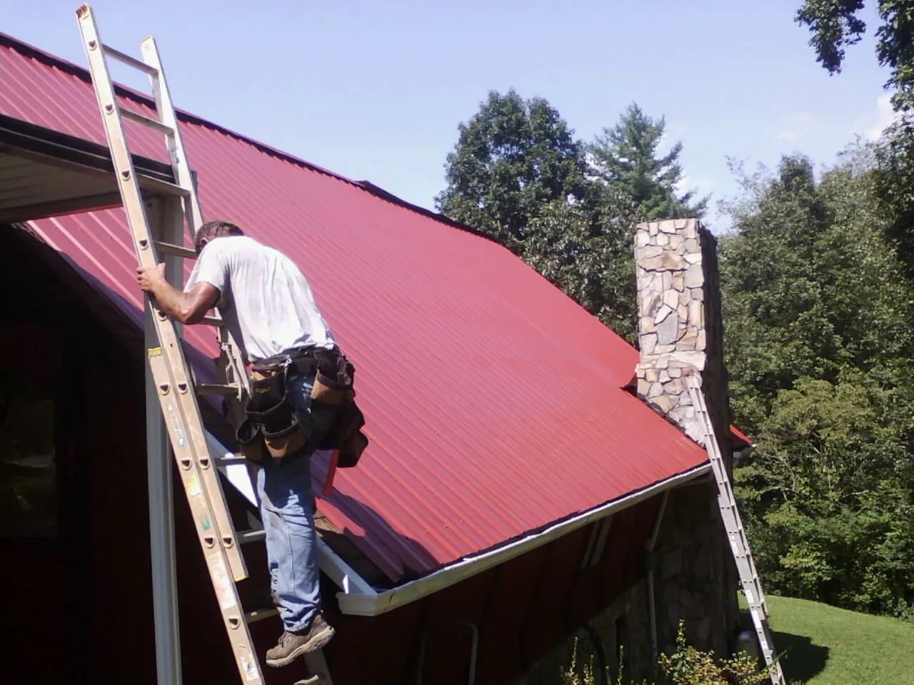 Roofer on a ladder beside a red metal roof and stone chimney — Soffit & Fascia Repair by Roofing Brothers in South Pekin